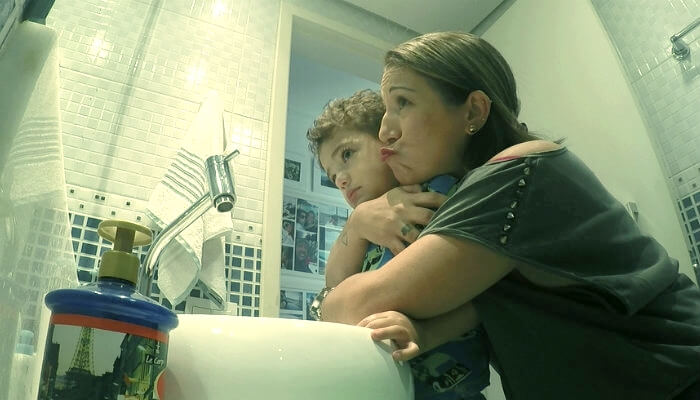 Elis, da Com Eira e Beira, em foto abraçando seu filho criança no banheiro. Os dois olham para o espelho