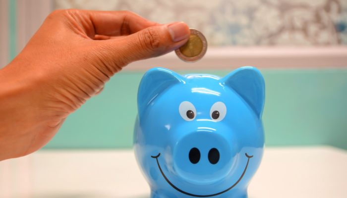 O que é e como aplicar a gestão financeira ao seu e-commerce? Cofre de porquinho azul com mão depositando uma moeda, representando a gestão financeira