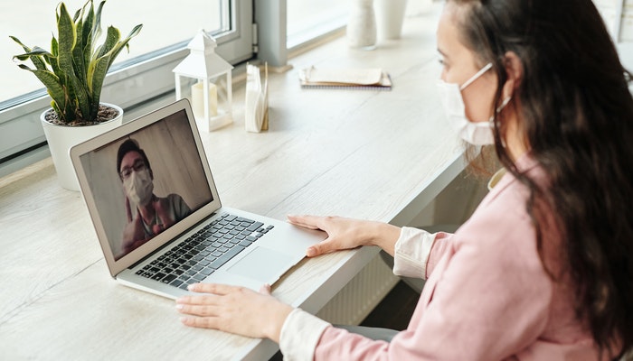 Responsabilidade social: como sua empresa pode ajudar no combate à Covid-19 Mulher de máscara em frente ao computador, em uma chamada de vídeo, representando o trabalho em uma empresa com responsabilidade social no combate à Covid-19