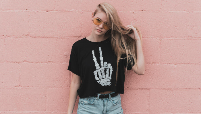 Loja de camisetas: o que considerar antes de montá-la? Modelo loira em frente a uma parede rosa olha para a câmera. Ela usa uma blusa preta com ilustração de ossos da mão fazendo símbolo de paz e amor, representando a loja de camisetas online