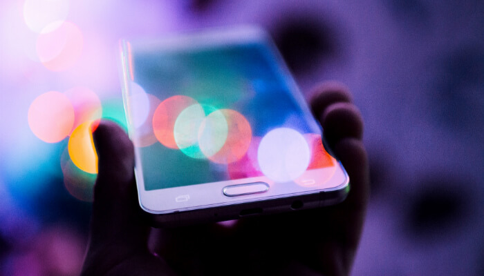 Mão segurando celular, que tem reflexos arroxeados na tela, representando o marketing nas redes sociais