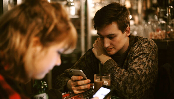 O poder do mobile-first index no seu marketing digital Homem e mulher sentados à mesa de um café, cada um mexendo em seu smartphone, representando o mobile first index