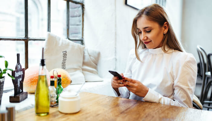 6 redes sociais para você considerar para seu e-commerce de moda Mulher com blusa branca estilosa sentada à mesa, mexendo no celular, representando as redes sociais para ecommerce de moda