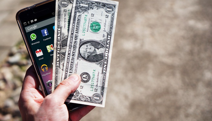Como criar sua loja virtual pelo aplicativo da Nuvemshop Mão segura smartphone e cédulas, representando o aplicativo da Nuvemshop