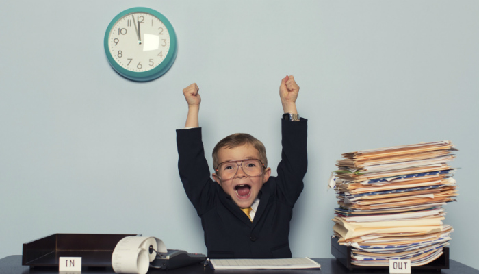 Como gerenciar as suas tarefas e aumentar a sua produtividade Menino com pose de comemoração em mesa de trabalho, representando como gerenciar tarefas