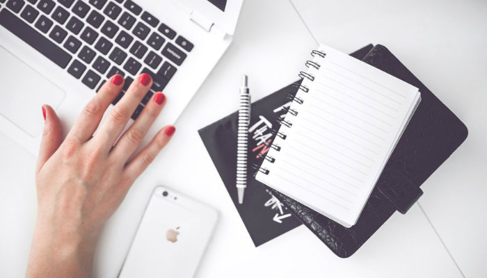 Como organizar as publicações em sua página do Facebook e aumentar sua audiência Mão de mulher em notebook, com smartphone e caderno ao lado, representa cronograma de postagens no Facebook