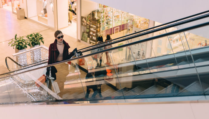 6 dicas para seu e-commerce de moda se diferenciar da concorrência Mulher com sacolas em escada rolante de shopping representa as dicas para loja virtual de roupas