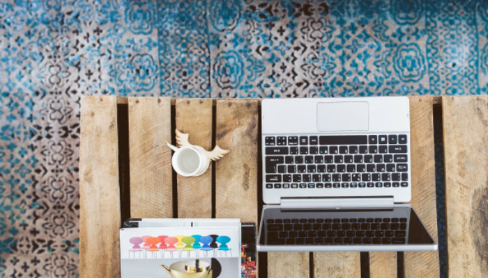 6 excelentes ferramentas sociais para quem trabalha remotamente Notebook sobre mesa de madeira com chão de ladrilhos coloridos ao fundo representa o trabalho remoto
