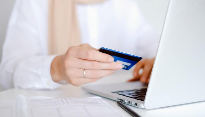 Imagem de uma mulher segurando um cartão de crédito em frente a um computador. Representando uma compra online.