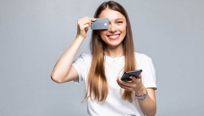 Na imagem, uma mulher branca e de cabelos claros segura um cartão de crédito na mão direita em frente aos olhos e um smartphone na mão esquerda.