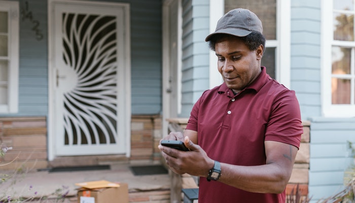 Na imagem, um homem negro de boina está mexendo no celular em frente a uma residência.