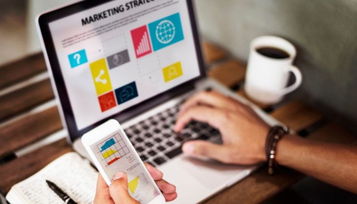 Saiba por que investir em marketing digital é importante para o seu negócio homem segurando um celular e traçando estratégias de marca em um computador, ressaltando a importância de investir em marketing digital
