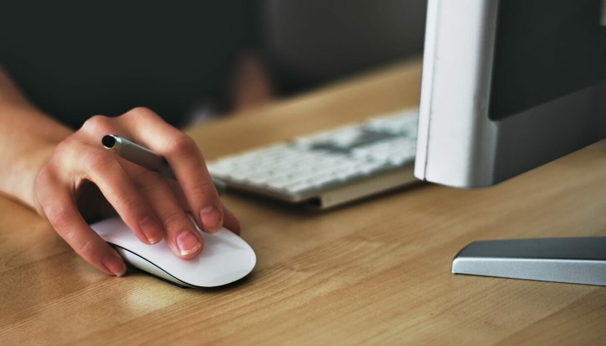 Mão sobre mouse, ao lado de teclado e computador, representando o conteúdo interativo