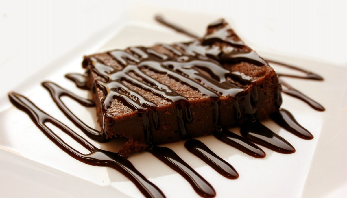 Na imagem, vemos uma fatia de brownie com cobertura de chocolate sobre um prato
