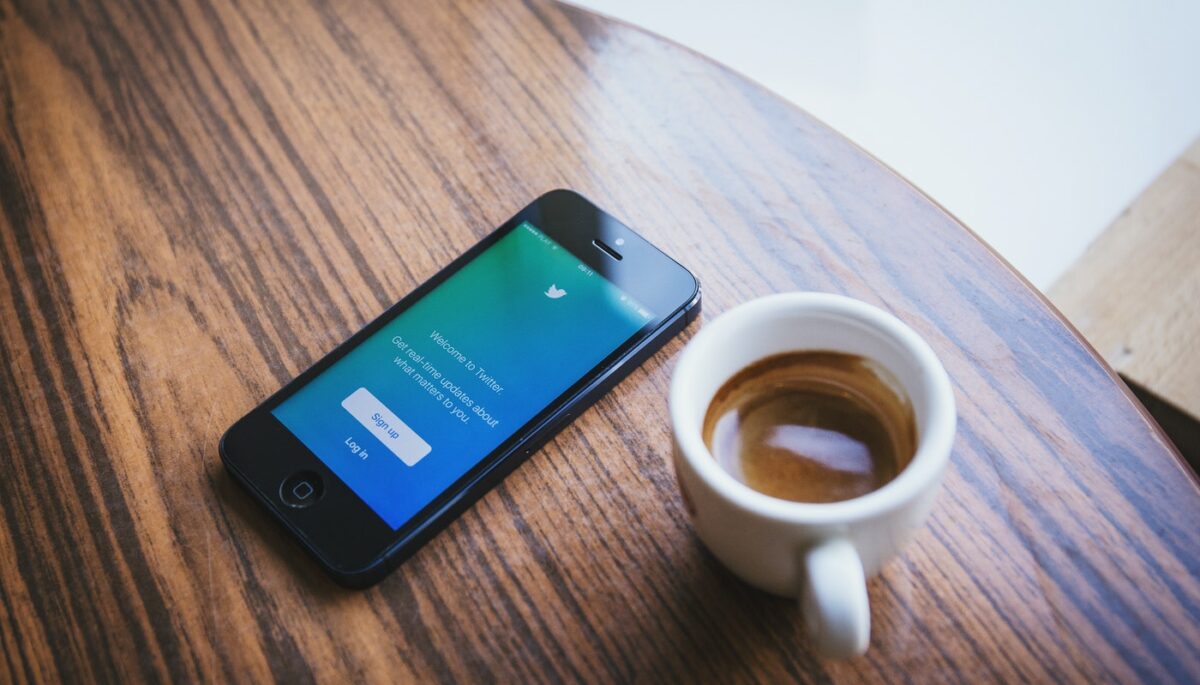 Como vender pelo Twitter e aumentar o reconhecimento da minha marca? Na imagem, tem um smartphone e uma caneca com café em uma mesa de madeira