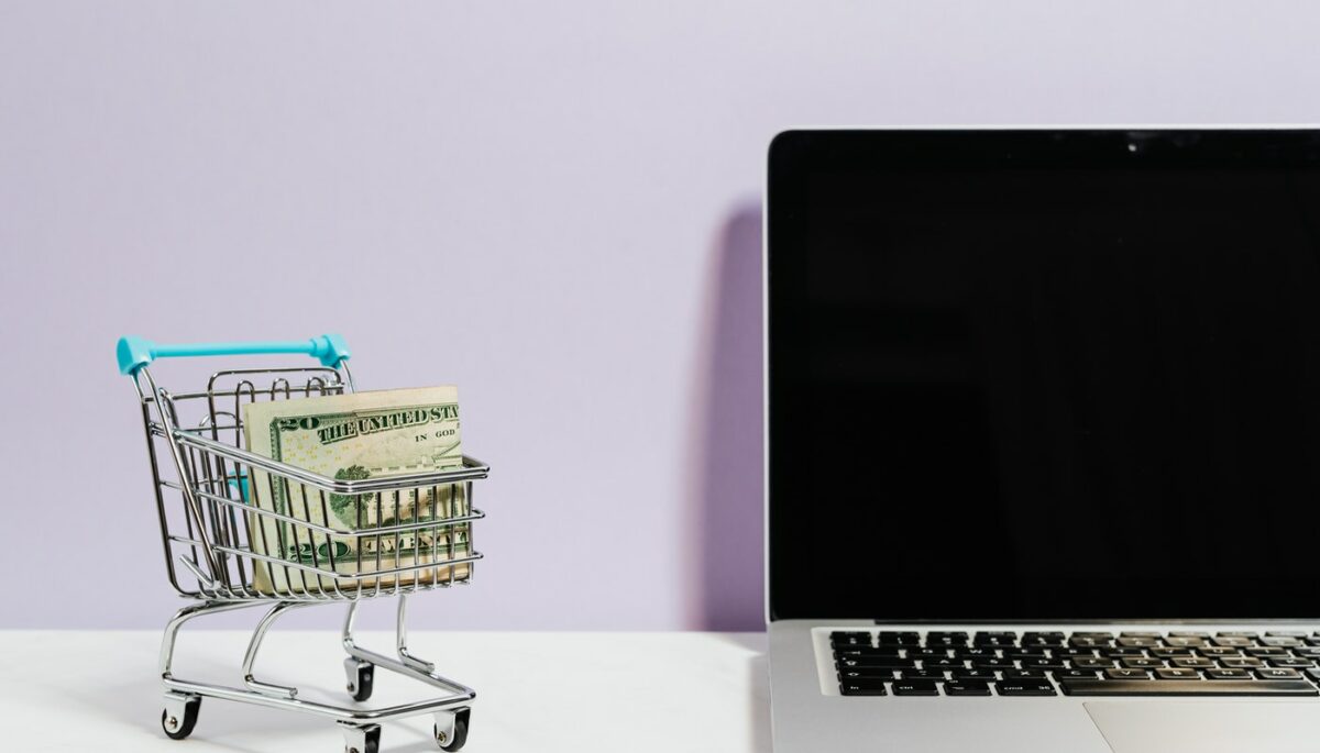 Carrinho de loja com dinheiro dentro ao lado de notebook, simbolizando a ideia de como montar uma loja virtual com pouco dinheiro