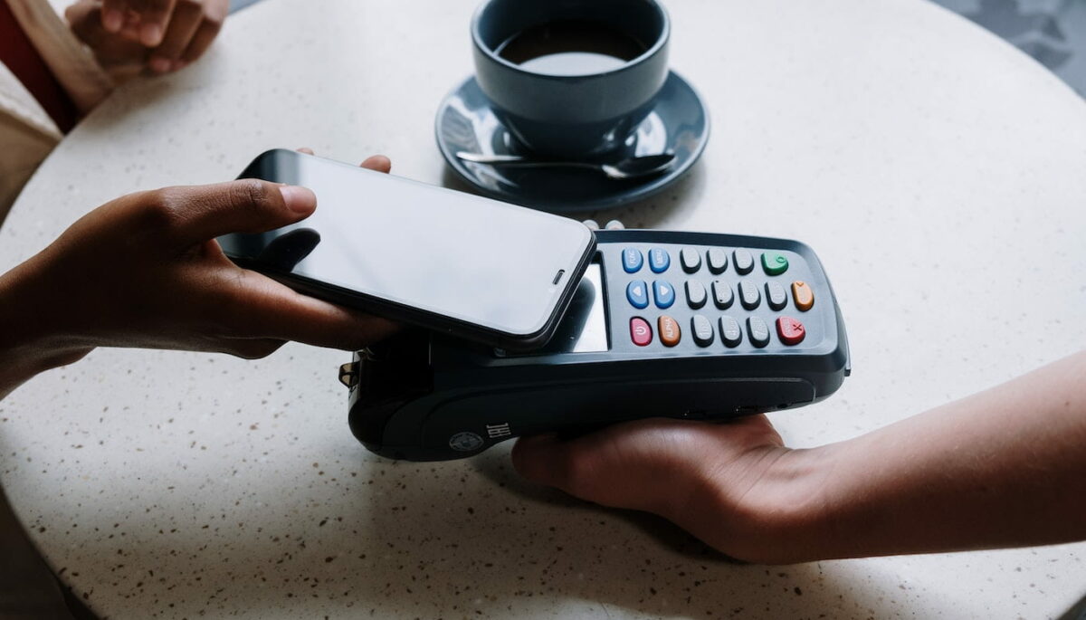 Mão aproximando celular de maquininha de cartão, como quem está explorando as diferentes opções de meios de pagamento eletrônicos