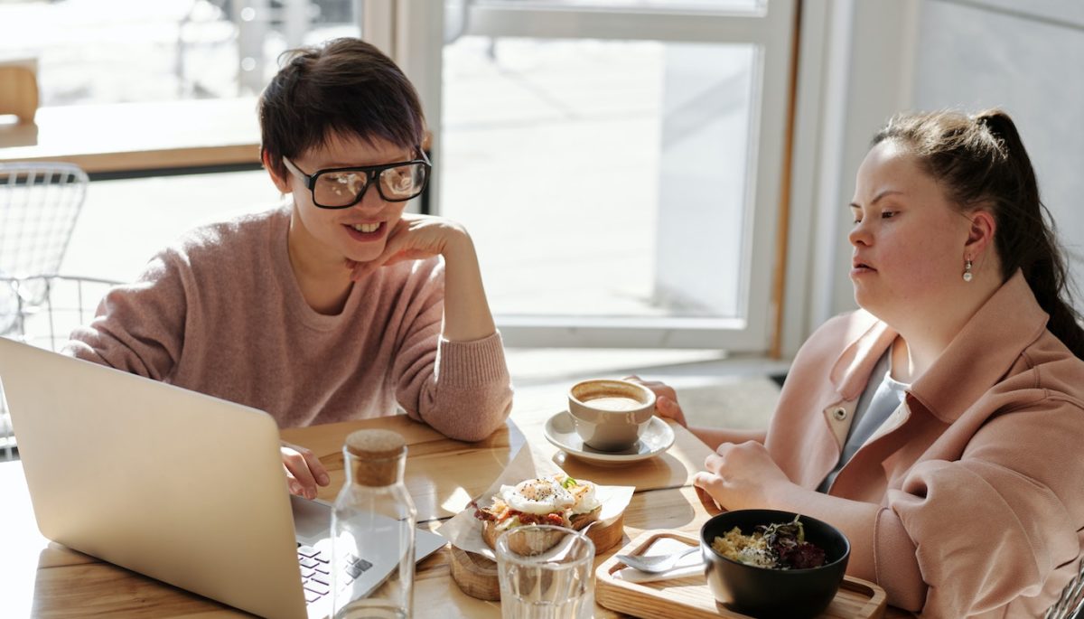 Na imagem, vemos duas amigas conversando e tomando um café com um notebook aberto na mesa.