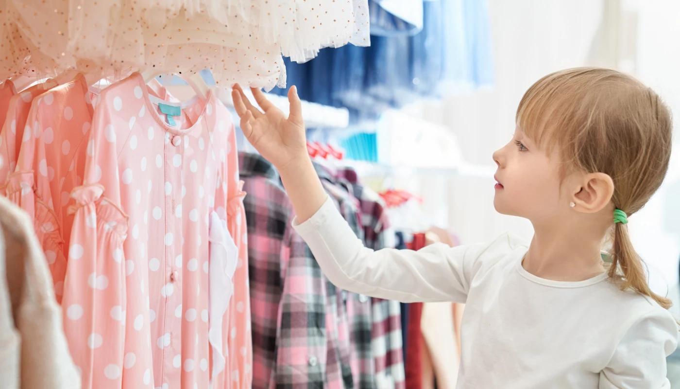 Criança menina toca peças de roupa infantis penduradas em araras de loja. Imagem faz referência à campanha de Dia das Crianças.