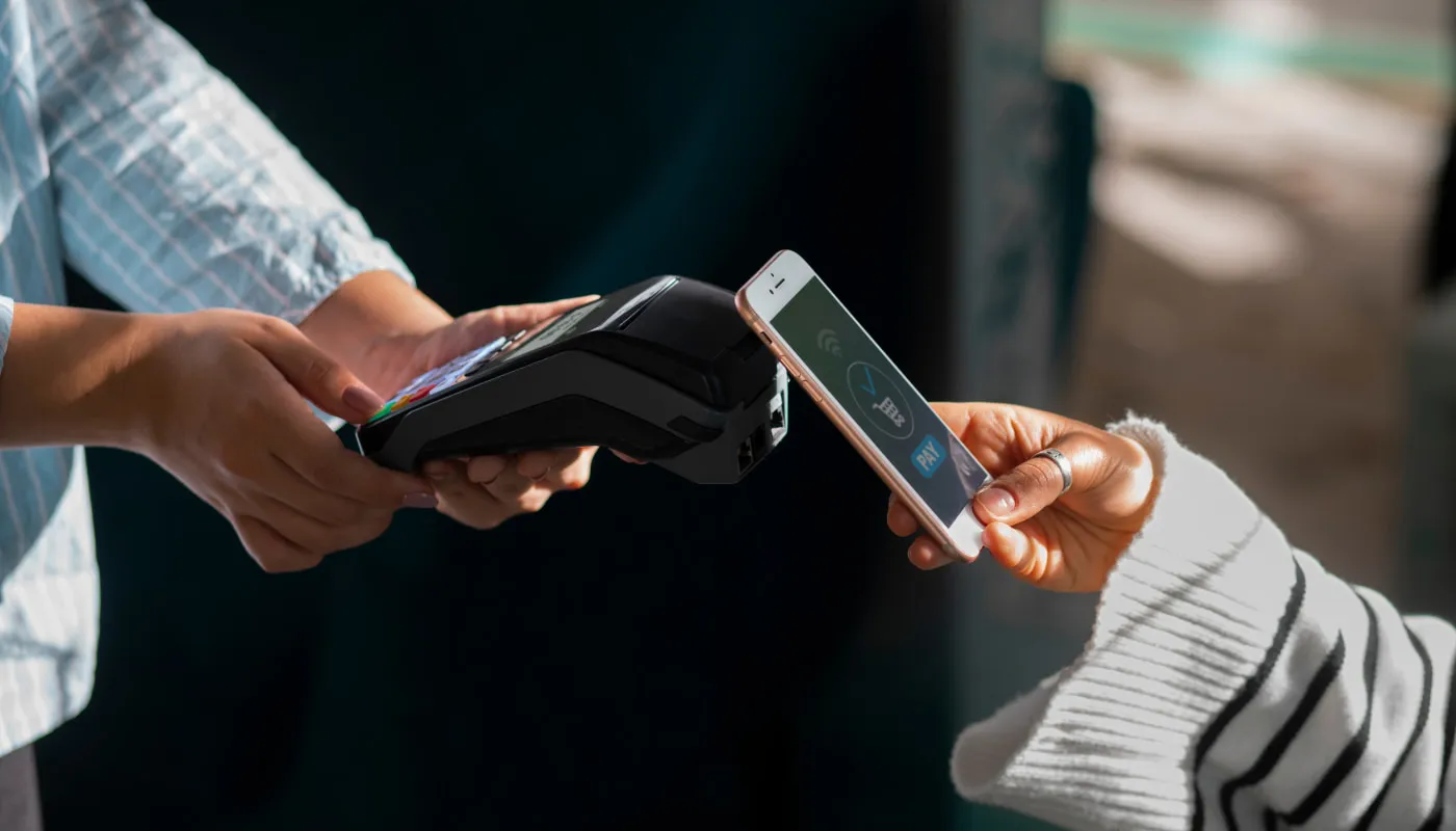 Na imagem, vemos as mãos de uma pessoa segurando uma máquina de cartão e outra pessoa finalizando a compra aproximando o celular, representando a nova moeda digital Drex.