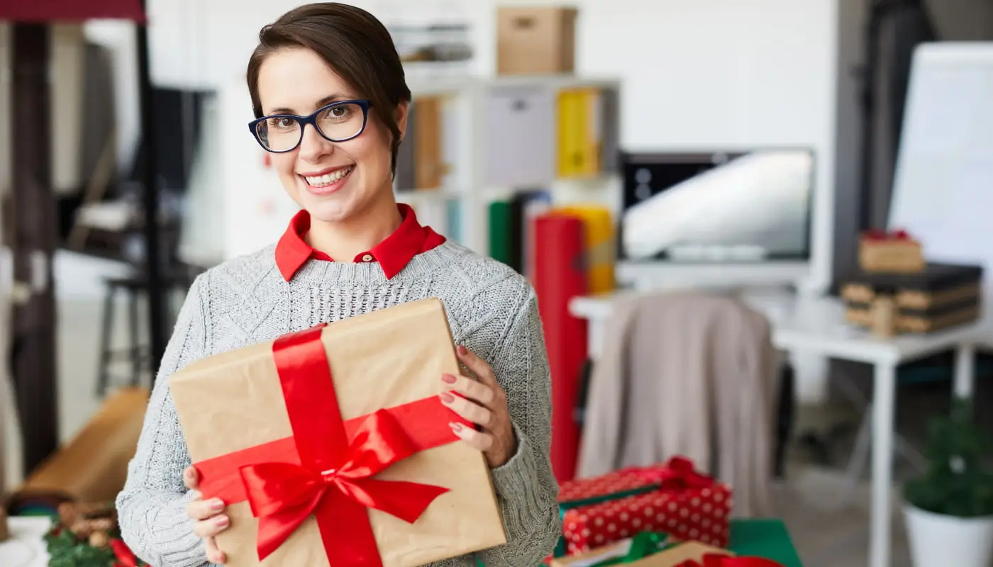 Na imagem, vemos uma mulher de óculos e cabelos curtos segurando um pacote de presente com laço vermelho em referência às vendas de natal.