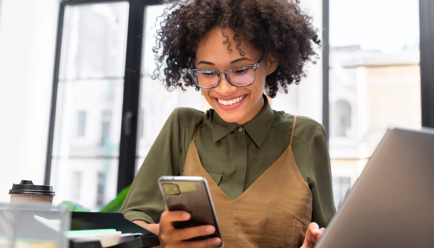 Mulher negra de cabelos cacheados sorri para o celular. Ela está de avental, indicando ser uma empreendedora pesquisando como abrir uma empresa.