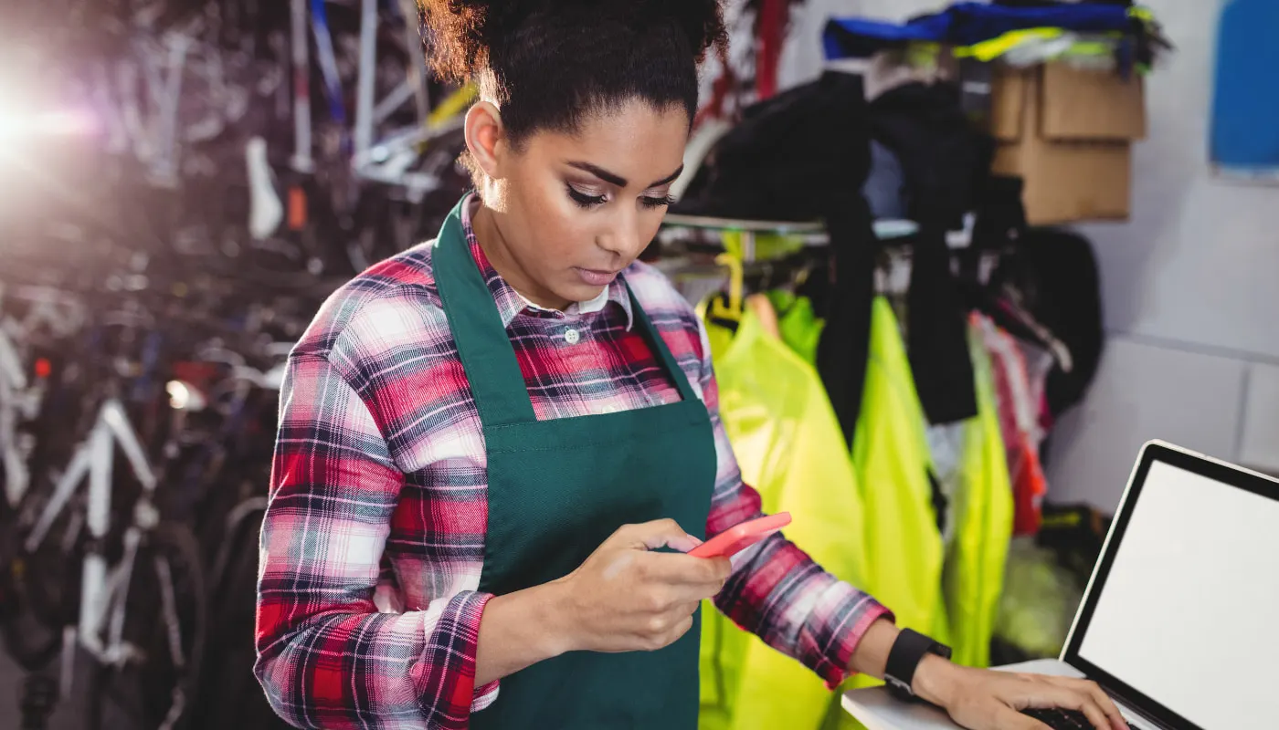 Mulher negra, com os cabelos presos e usando um avental, está em uma loja mexendo no telefone. Imagem faz referência ao uso do app Astroselling.