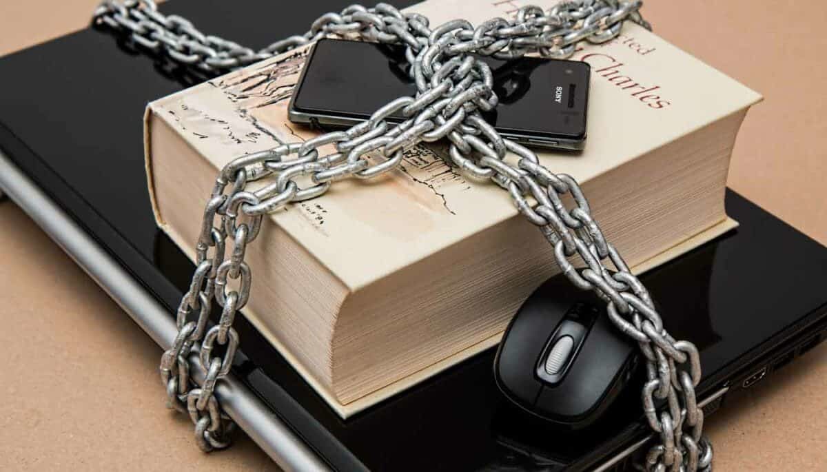 Computador com livro, mouse e smartphone amarrados com uma corrente representam como vender pela internet com segurança.