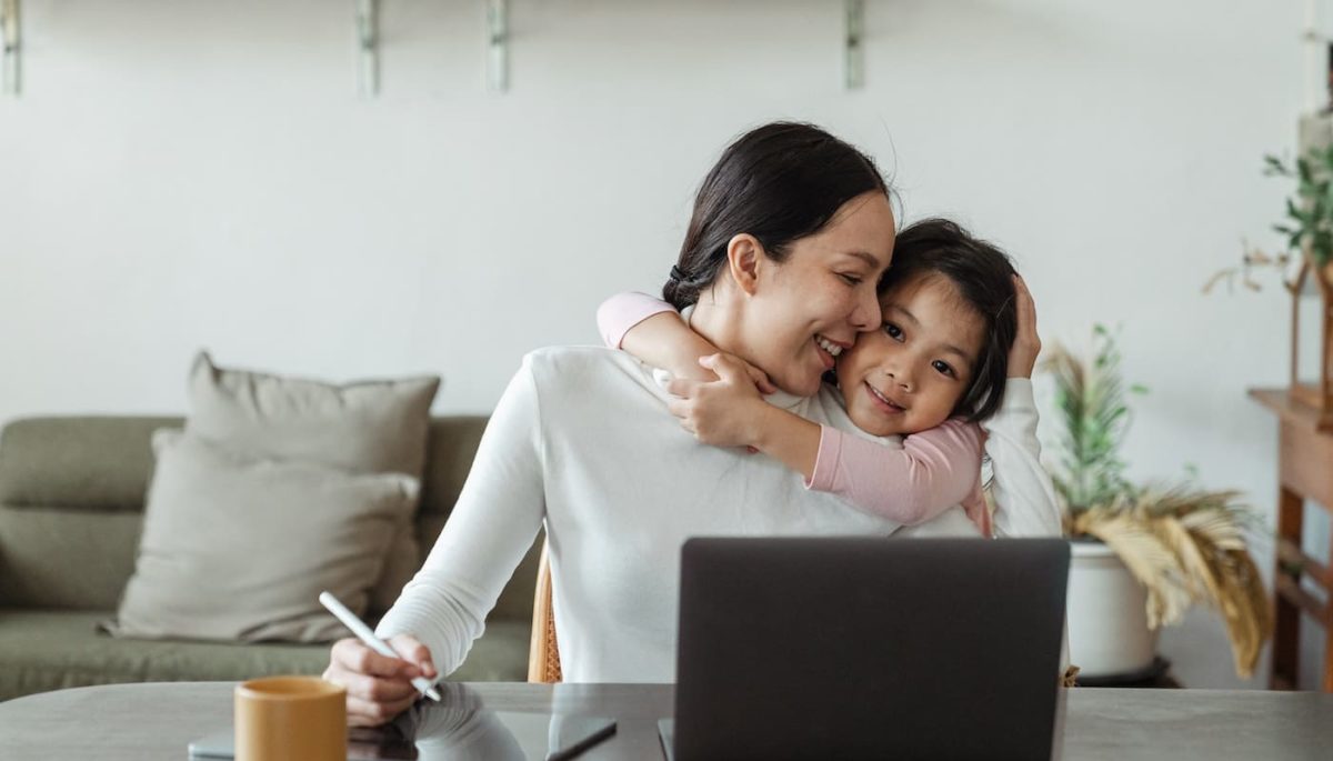 Mulher em frente ao computador e ao lado de uma criança, representando o que é ser mãe empreendedora.