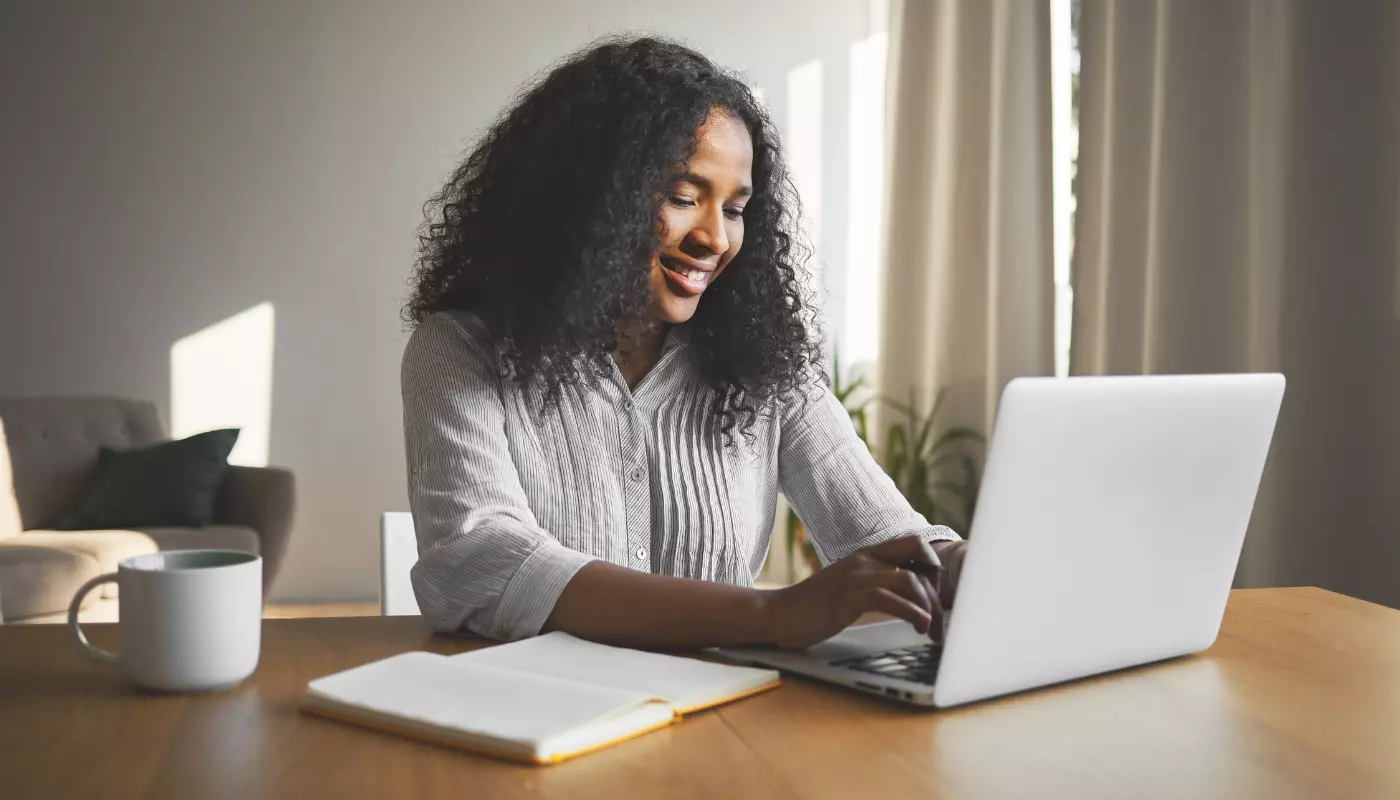 Imagem mostra uma mulher negra de cabelos soltos sentada de frente para um notebook e com um caderno ao lado planejando uma estratégia de marketing de conteúdo.