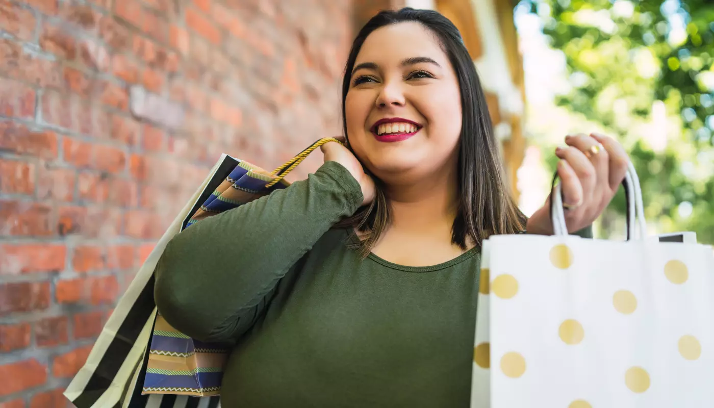 Mulher plus size branca de cabelos pretos e lisos aparece sorrindo e segurando várias sacolas. Imagem faz referência à saber como vender roupas plus size.