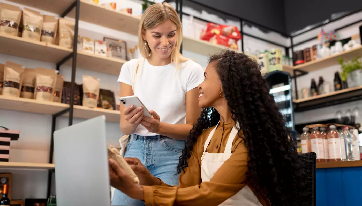 Imagem mostra uma mulher loira mostrando um produto no celular para uma mulher negra. Ao fundo, vemos um grande estoque de itens, representando os fornecedores de produtos para revenda.