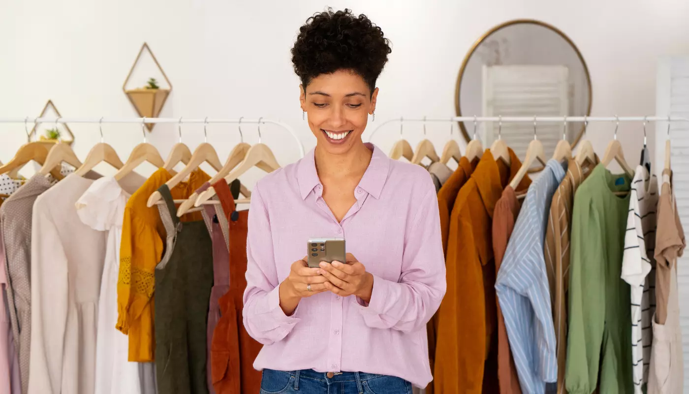 Mulher negra está na frente de uma arara de roupas sorrindo e olhando para o celular. Imagem faz referência a uma empreendedora escolhendo um nome para loja de roupas.