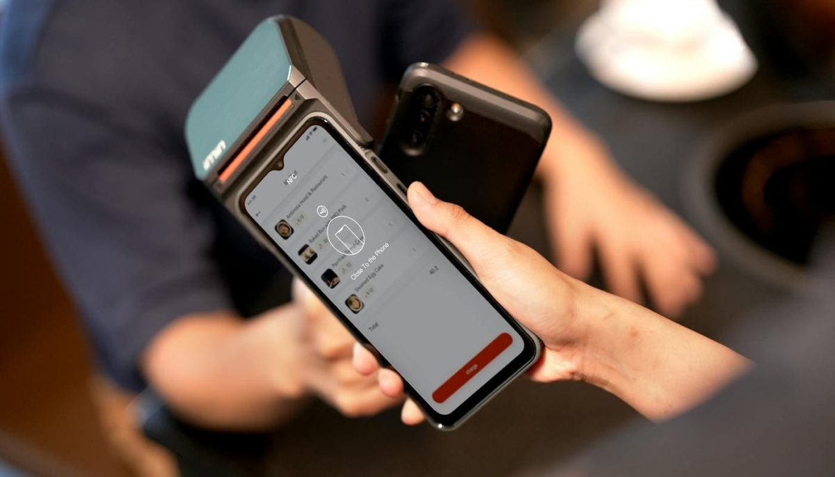 Imagem de pessoa aproximando o celular de uma maquininha de cartão, representando pagamento por cartão virtual.