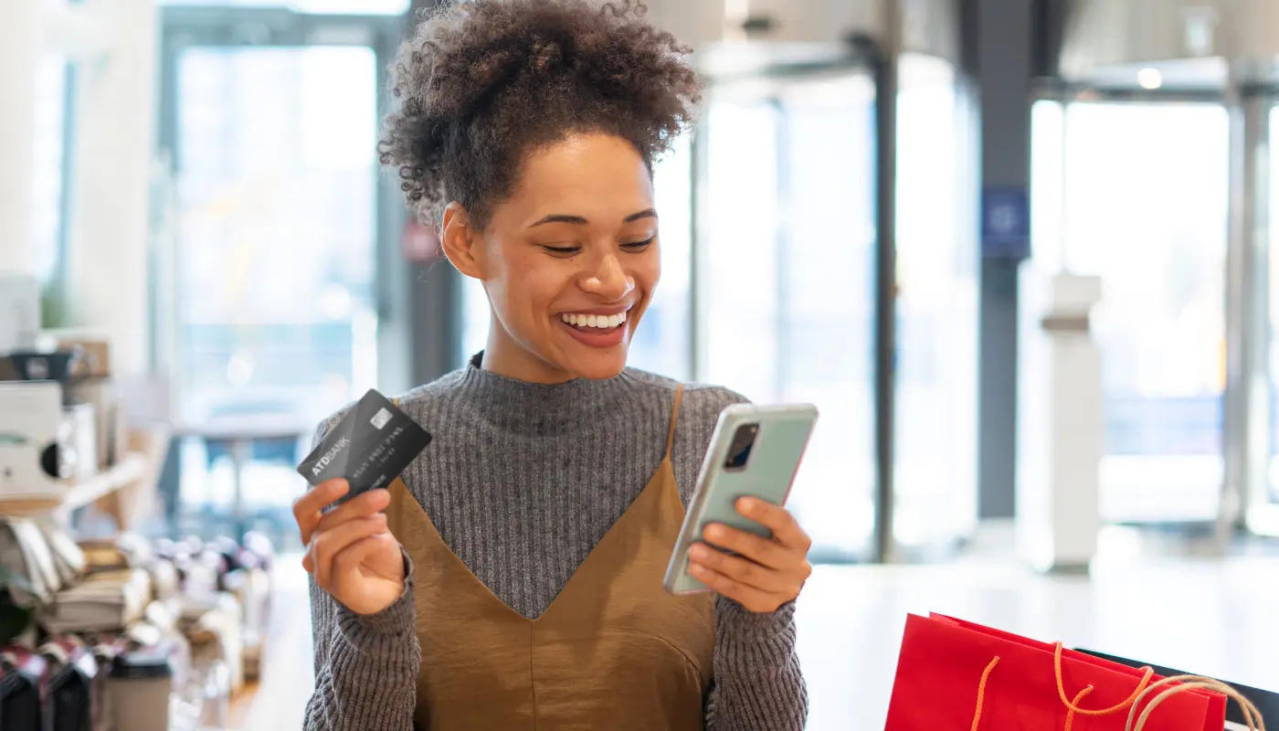Mulher negra está sorrindo enquanto olha para o smartphone em sua mão. Na outra mão, ela segura um cartão para fazer compras na Cyber Monday.