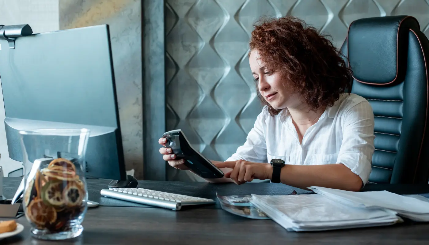 Empreendedora está sentada em seu escritório com uma calculadora nas mãos e pesquisando sobre a substituição tributária no computador à sua frente.