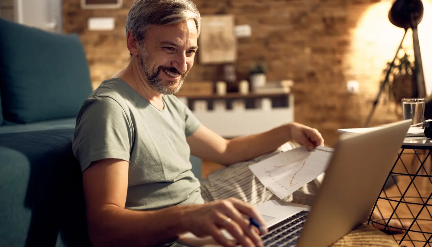 Homem de meia idade sentado no sofá com um notebook no colo e folhas com gráficos indicando como ganhar dinheiro na internet.