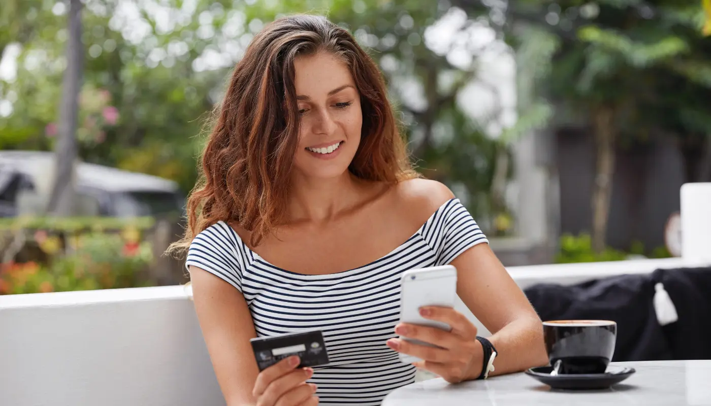 Mulher jovem segura o telefone em uma mão e o cartão de crédito na outra, usando o CVV para comprar online.
