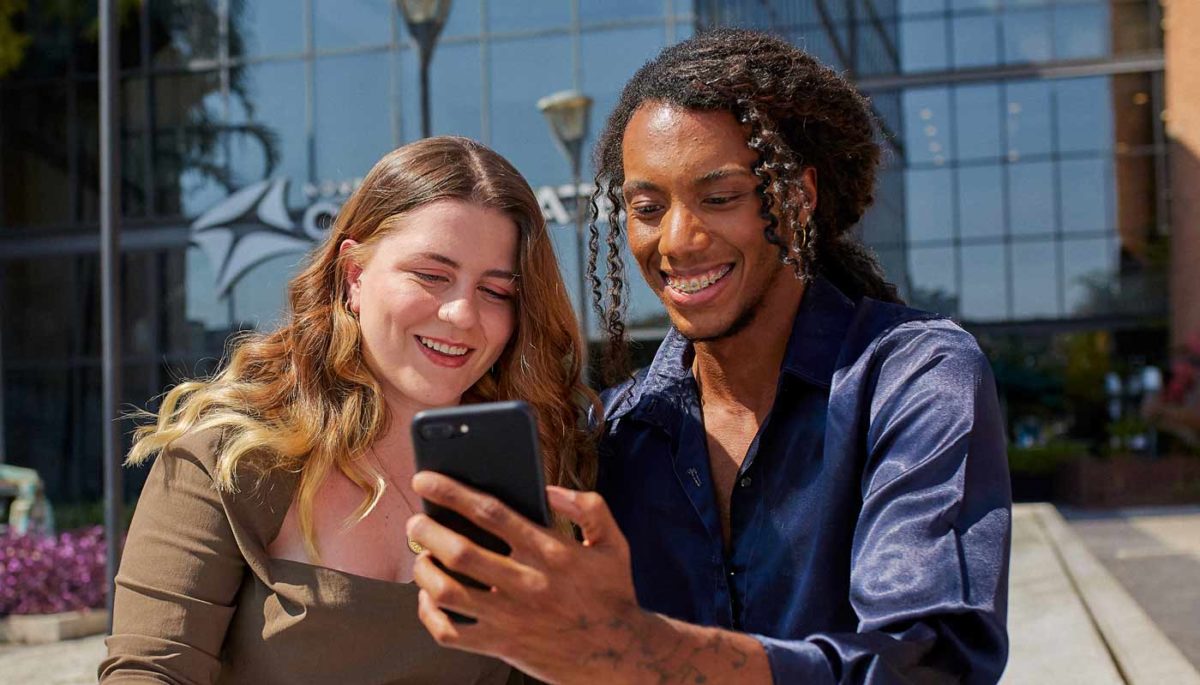 Rapaz negro de cabelos longos mostra os serviços financeiros que utiliza em seu aplicativo bancário para uma mulher branca. Ambos sorriem.