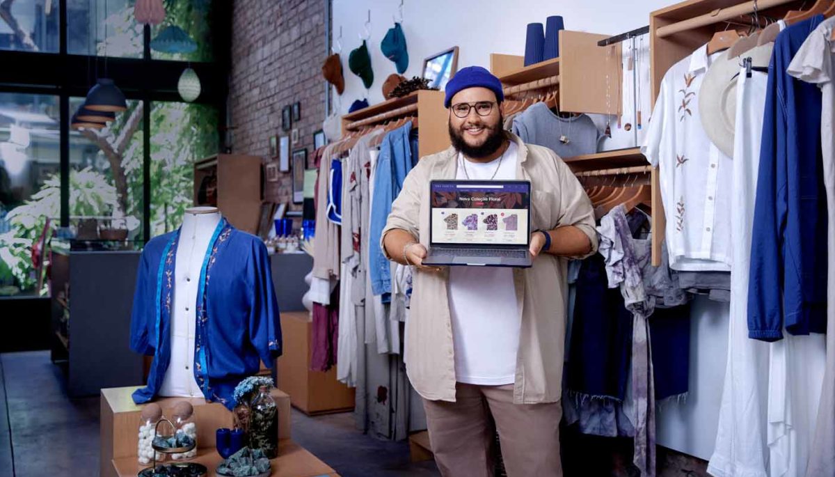 Empreendedor que possui B2C ecommerce na Nuvemshop mostra sua loja virtual em um notebook, com os produtos da loja de roupas ao fundo.