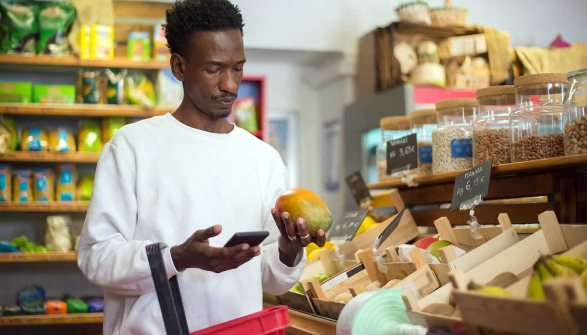 Consumidor negro faz compras na loja física enquanto pesquisa preços no smartphone, representando o digital commerce.