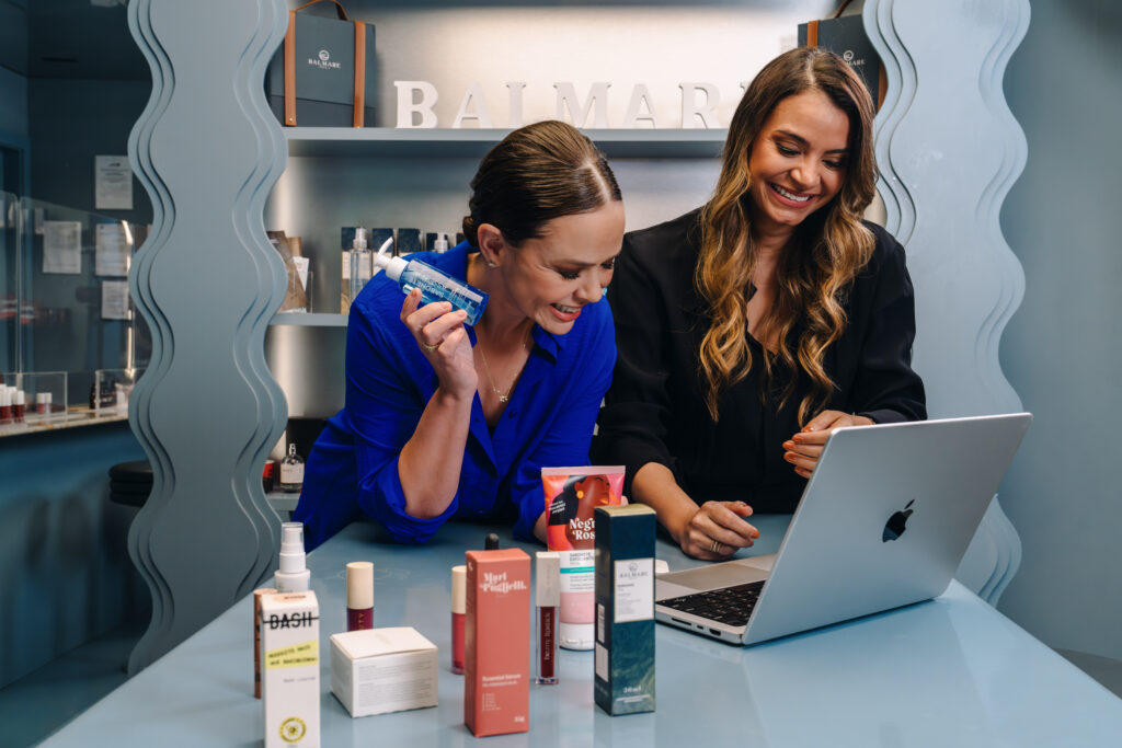 Imagem mostra Simone Sancho e Amanda Coelho, fundadoras da Belong Be, olhando para tela de computador com cosméticos da Belong Be ao redor
