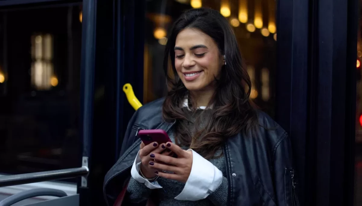 Buy Now Pay Later: como funciona o “compre agora, pague depois”? Uma mulher sorridente em um ônibus olha para o celular, considerando uma compra com a opção