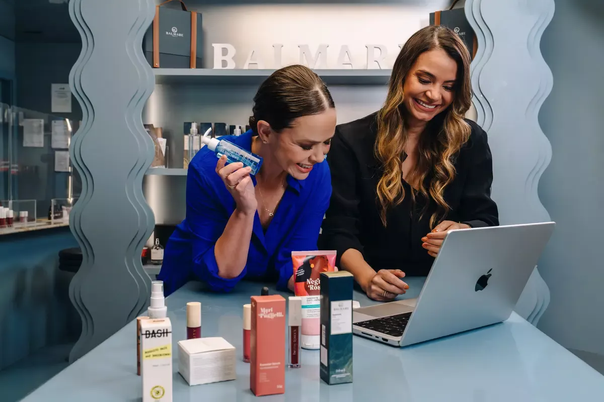Imagem mostra Simone Sancho e Amanda Coelho, fundadoras da Belong Be, olhando para tela de computador com cosméticos da Belong Be ao redor