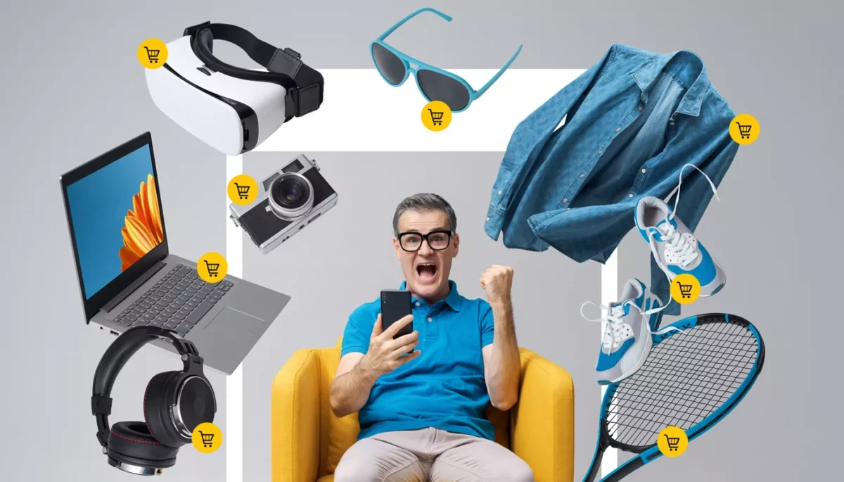 Um homem, representando o consumidor digital, sentado em uma poltrona amarela, expressa excitação enquanto segura um smartphone, cercado por diversos itens flutuantes como óculos VR, óculos de sol, uma câmera, um laptop, fones de ouvido, uma camisa jeans, tênis e uma raquete de tênis, todos com ícones de carrinho de compras, ilustrando a variedade de produtos disponíveis para compra online.